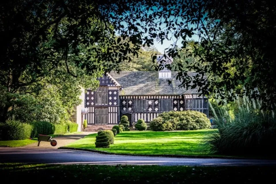 Rufford Old Hall's Tudor timbered façade reflected in a still ornamental pond, framed by late-summer greenery.