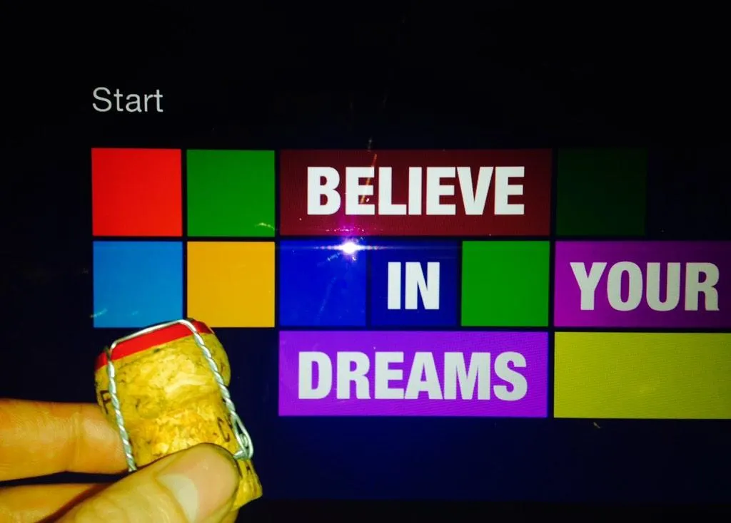 A 'Believe in your dreams' message laid out as colourful Windows Start-screen tiles, with a hand holding a champagne cork in front of the screen.