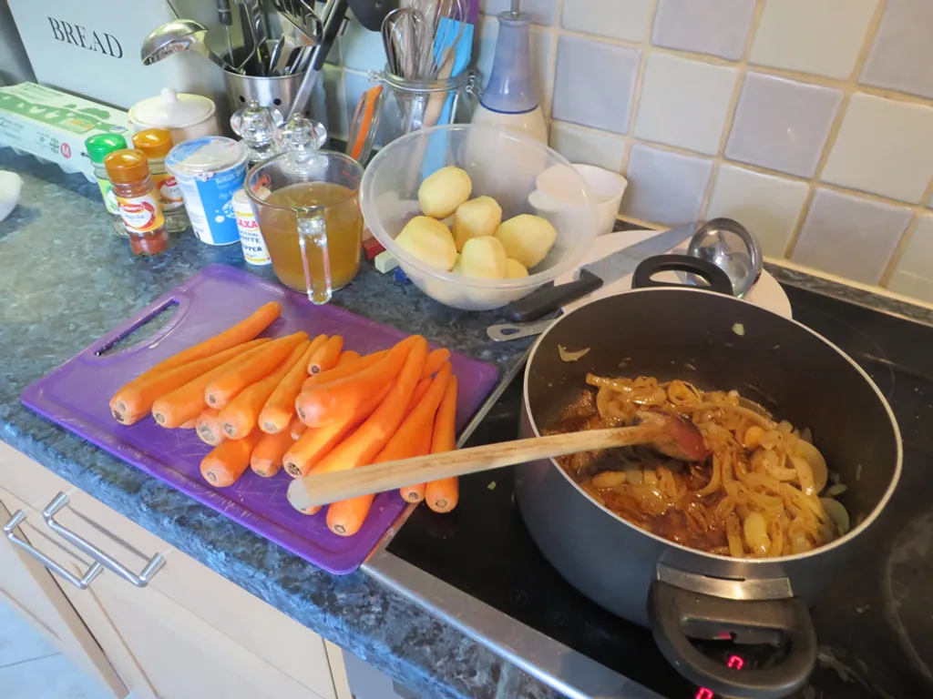 Kitchen worktop with peeled carrots on a chopping board, peeled potatoes in a bowl, a measuring jug of vegetable stock, paprika, white pepper, and a saucepan of sweating onions on the hob.