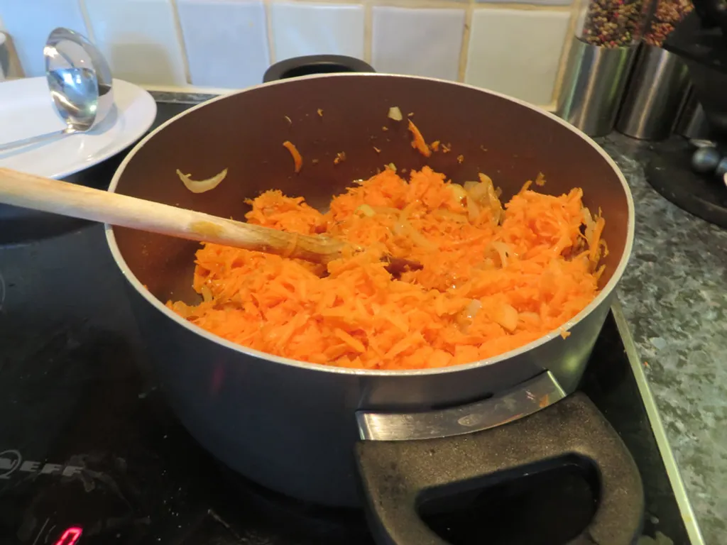 A saucepan on the hob with finely grated orange carrots piled on top of softened onions, a wooden spoon resting in the pan.