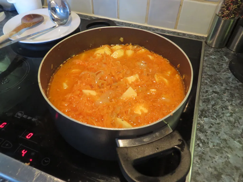 A saucepan simmering on a hob, full of orange carrot and potato in a translucent broth with onion strands visible.