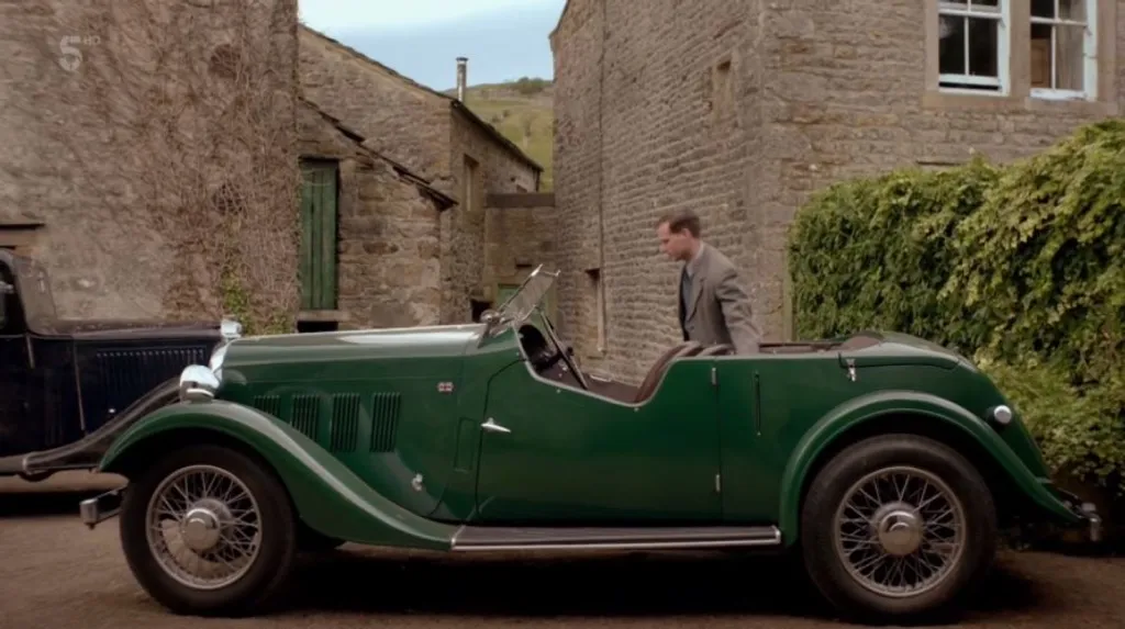 A gleaming dark green vintage open-top sports car parked in a narrow Yorkshire stone courtyard, a man in a period grey suit stepping into the driver's seat.