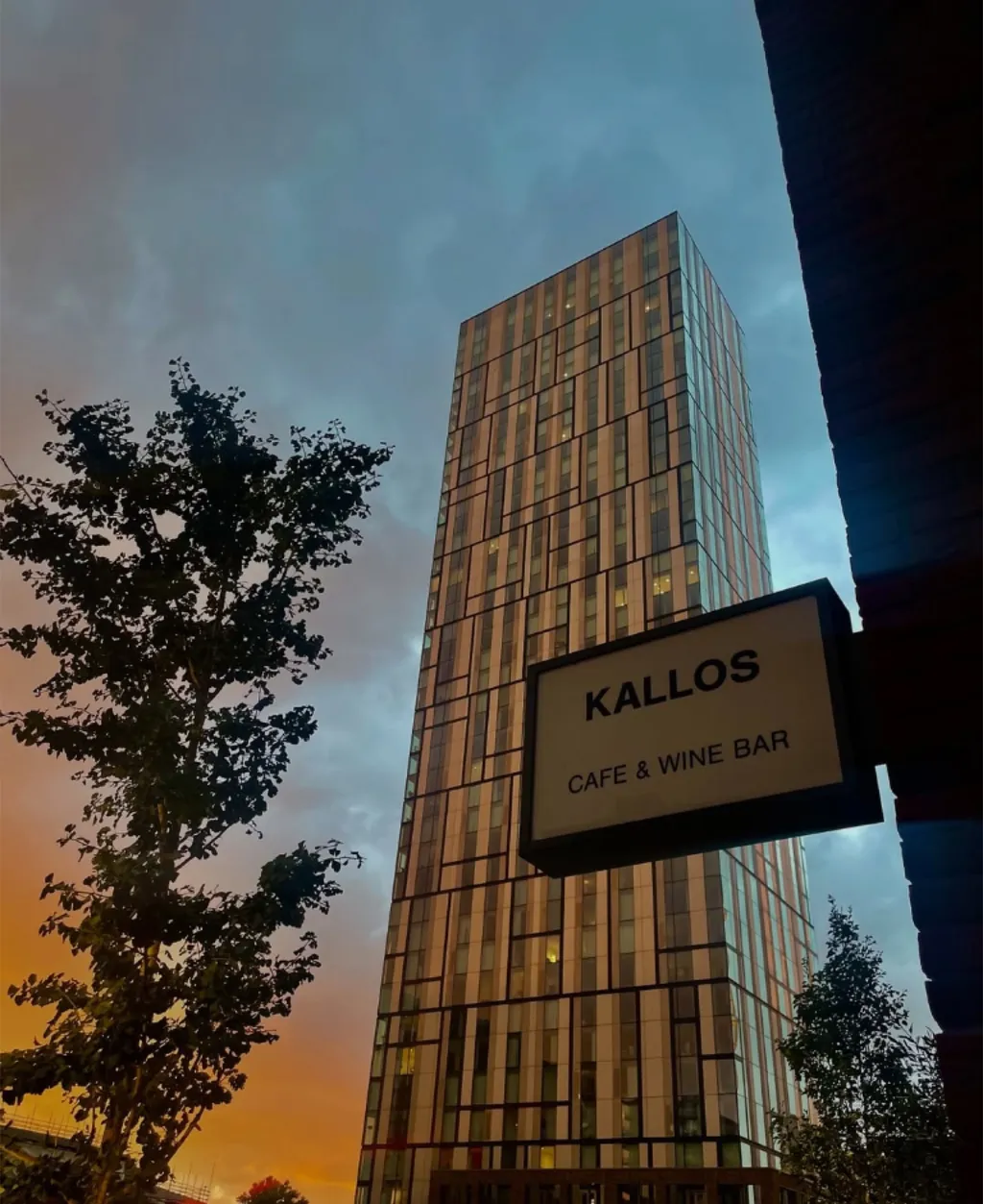 The exterior of Kallos Cafe & Wine Bar at dusk, the blue Kallos sign glowing beside the warm-lit windows, an illuminated tower behind.