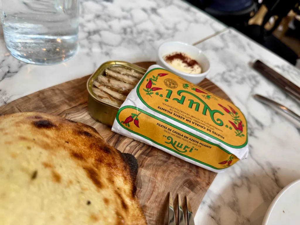 An open Nuri tin of spiced mackerel fillets in olive oil on a wooden board, the yellow and green wrapper folded back, with a small dish of paprika-dusted aioli behind it and the edge of a charred pita in the foreground.