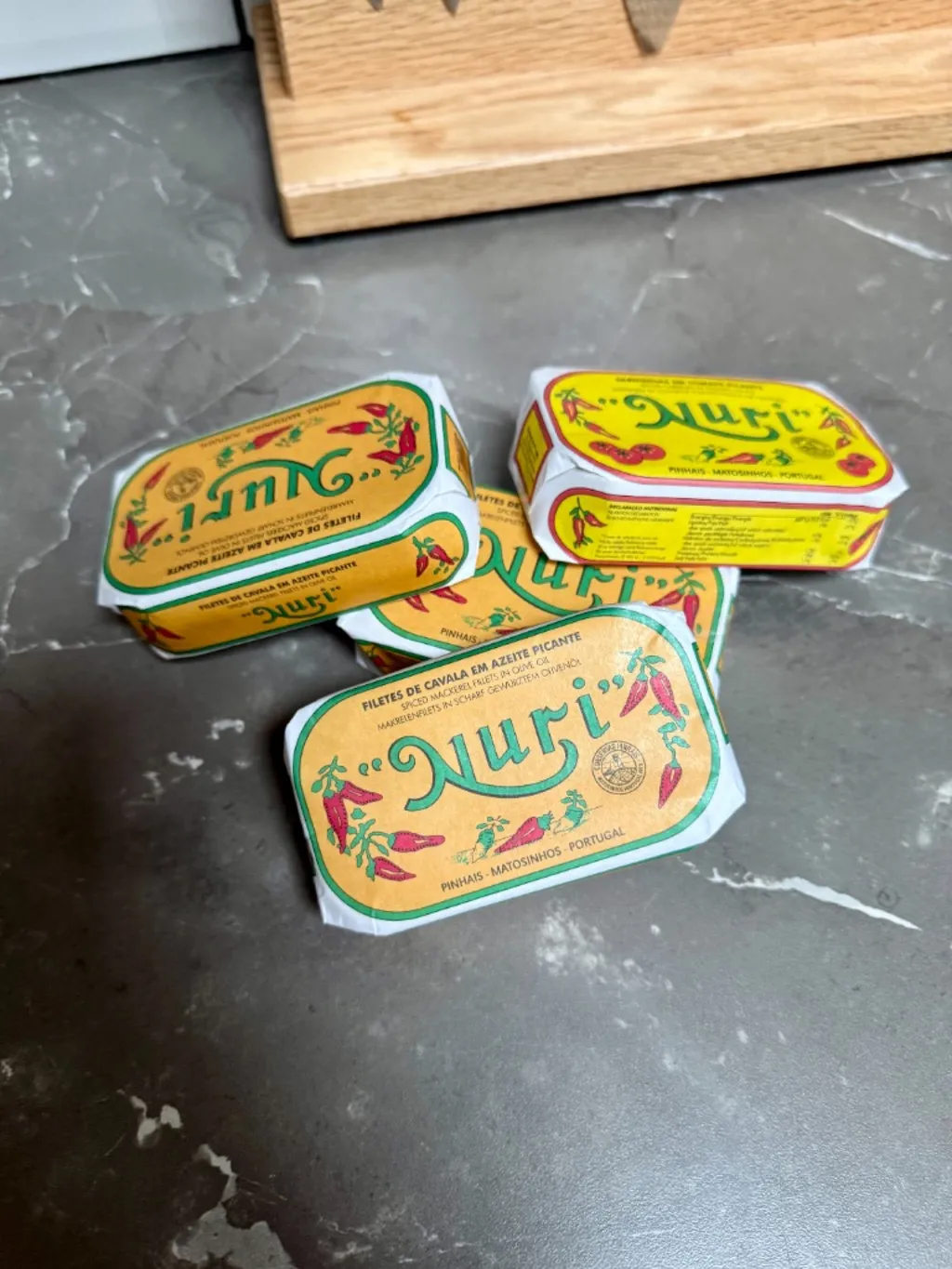 Three Nuri tins of spiced mackerel fillets in olive oil arranged on a grey marble counter in front of a wooden chopping board, their yellow paper wrappers showing the green 'Nuri' logo, red chilli pepper illustrations, and 'Pinhais, Matosinhos, Portugal' along the lower edge.