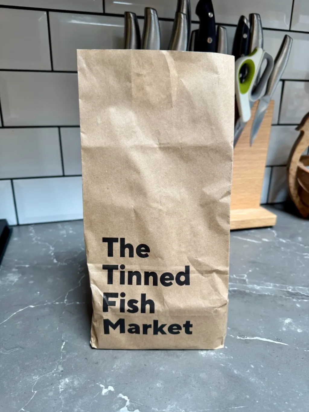 A brown kraft paper grocery bag printed with 'The Tinned Fish Market' in bold black sans-serif, standing on a grey marble worktop in front of a knife block, against a white metro-tiled wall.