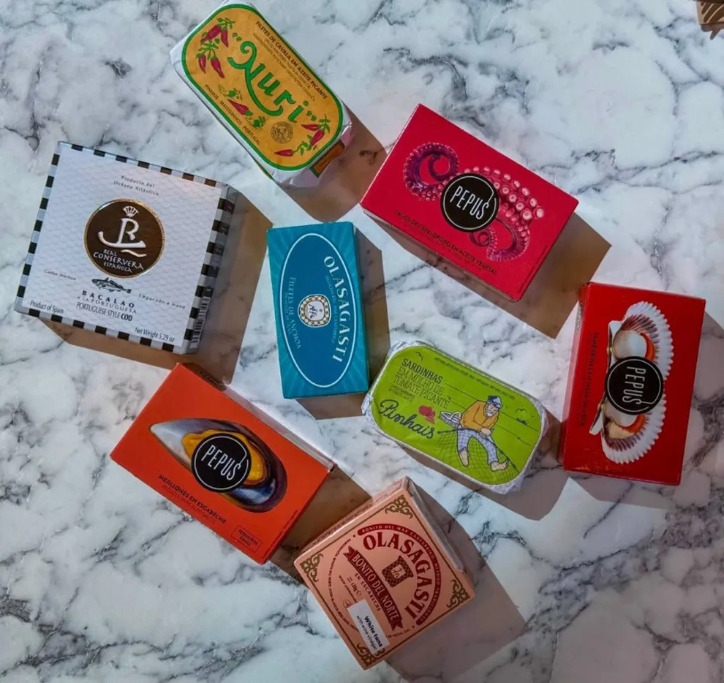 A flat-lay of premium tinned fish boxes on the marble table at Kallos: Nuri, Pinhais, Pepus, Olasagasti and others, stacked alongside a pita and small dishes.