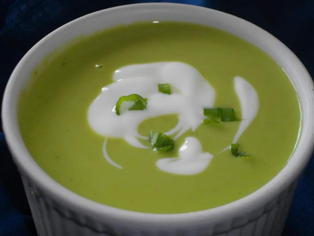 A white bowl of vivid green pea soup with a swirl of cream and a sprinkle of chives, on a wooden surface.