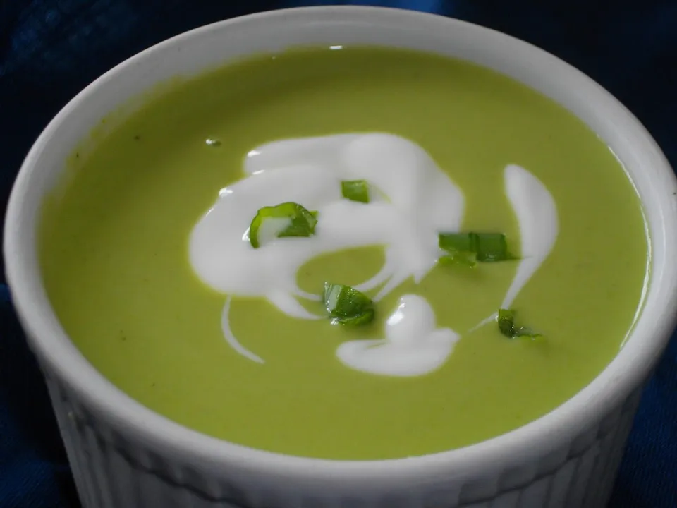 A white bowl of vivid green pea soup with a swirl of cream and a sprinkle of chives, photographed from above on a wooden surface.