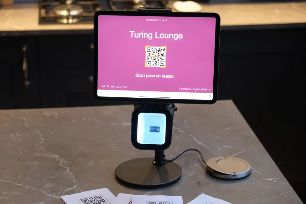 A first prototype of the Power App entry kiosk running on an iPad propped up on a wooden surface, showing the entry-point screen with a barcode scanner cable trailing out of frame.